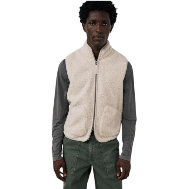 A man is wearing the HARTFORD - BASTA SHERPA VEST IN NATURAL over a gray long-sleeve shirt, holding the vest’s zipper with one hand. The plain, light-colored background complements the cozy, Hartford-inspired style of this men’s vest from HARTFORD.