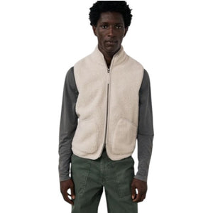 A man is wearing the HARTFORD - BASTA SHERPA VEST IN NATURAL over a gray long-sleeve shirt, holding the vest’s zipper with one hand. The plain, light-colored background complements the cozy, Hartford-inspired style of this men’s vest from HARTFORD.