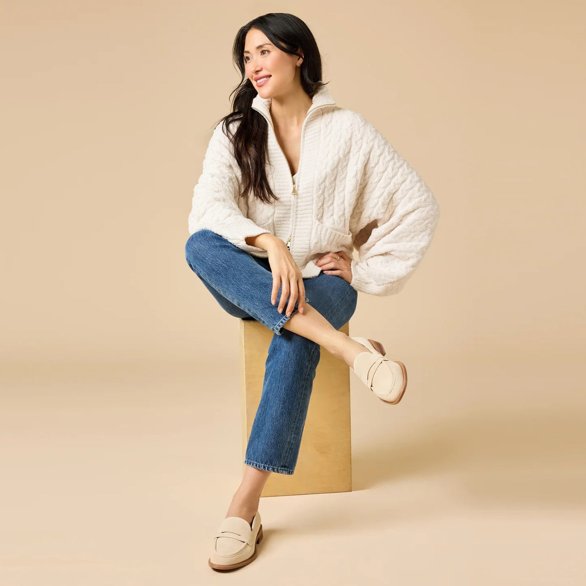 A woman wearing a cream-colored, plush SOFTIES - CABLE KNIT MARSHMALLOW FUNNEL NECK DOUBLE ZIP JACKET with a funnel neck and double zipper, paired with blue jeans.