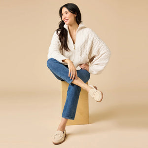 A woman wearing a cream-colored, plush SOFTIES - CABLE KNIT MARSHMALLOW FUNNEL NECK DOUBLE ZIP JACKET with a funnel neck and double zipper, paired with blue jeans.