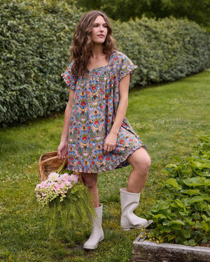 A woman with long blonde hair wears the PRINTFRESH Bee Tapestry Pinktuck Nightgown in Delphinium Blue, standing against a plain light-colored wall and smiling slightly.