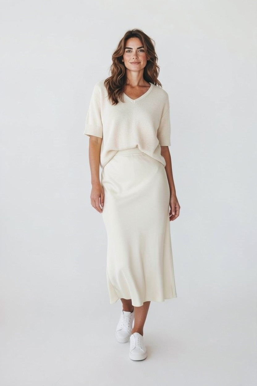 A woman with long wavy hair smiles softly against a light background, wearing the ROSE &amp; CROWN Cashmere V Neck Short Sleeve Sweater and a matching midi skirt, paired with white sneakers.