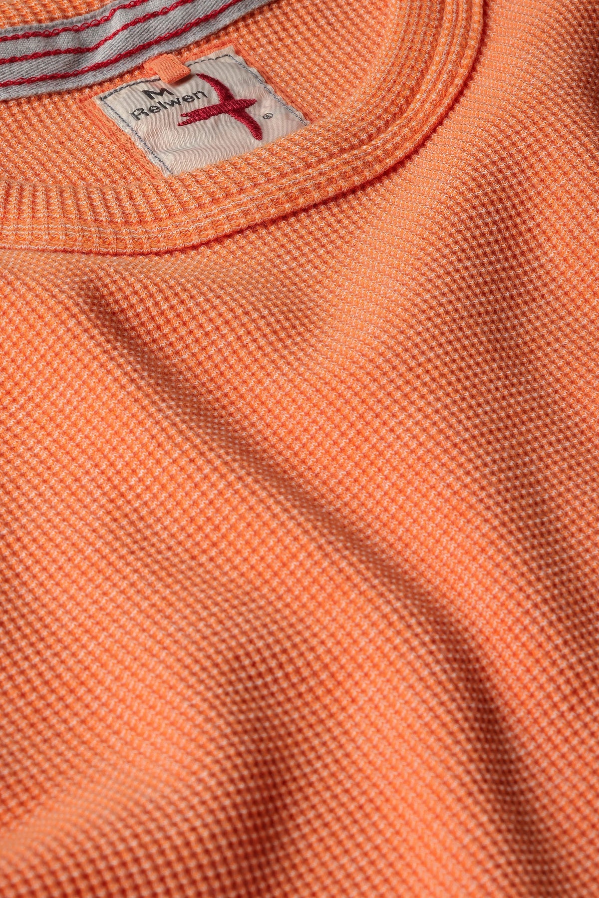 The RELWEN - BI-WAFFLE CREW IN CITRUS, a long-sleeved, citrus orange waffle-knit cotton sweater from RELWEN, is laid flat on a white background. It features ribbed cuffs, hem, and collar with a small visible fabric tag near the hem.