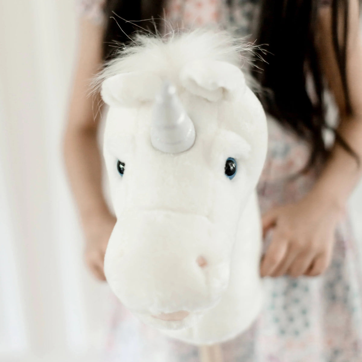 A young child in a cozy, bohemian-style bedroom holds the LITTLE UNICORN RIDER by LILY &amp; RIVER, sparking their imagination. The room showcases children's products such as a small wooden bed with a canopy, a table and chairs, and wall decorations made of macrame. Dressed in brown with hair styled in two buns, the child enjoys playful physical activity.