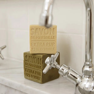 Two cubes of COMPAGNIE DE PROVENCE AUTHENTIC CUBE OF SOAP OLIVE OIL 13.8OZ by CIE LUXE BRANDS, one beige and one olive green, are stacked on a marble sink beside a chrome faucet. Each is labeled “Extra Pur 72% d’Huile.”.