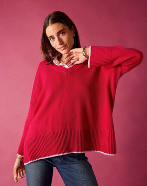 Wearing the MERSEA Montauk Colorblock V Neck Sweater in Raspberry, a woman in jeans poses against a pink background, smiling softly and raising one arm near her face.
