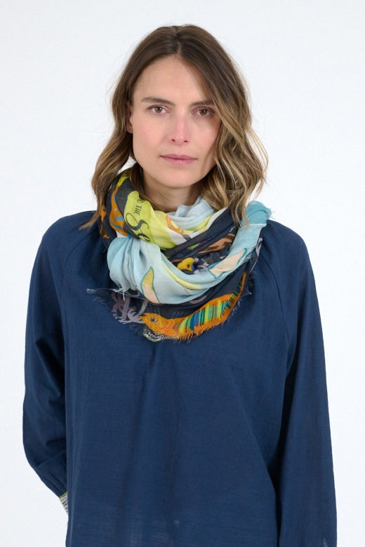 A woman with shoulder-length wavy hair models the STORIATIPIC - GERTRUD SCARF, styled around her neck over a navy blue top, against a plain white background.