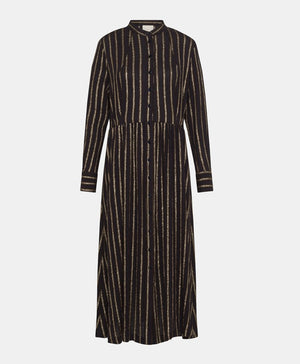A woman in the MOMONI Aubert Dress in Lurex Gauze stands by a woven chair, gazing down as her hair partially covers her face.