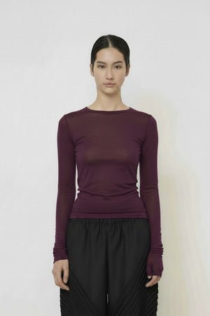 A woman with long dark hair in a low ponytail stands in profile against a plain light background, wearing the MEIMEIJ MESH LAYERING TOP by MEIMEIJ in red and a black skirt for a minimalist Italian fashion look.