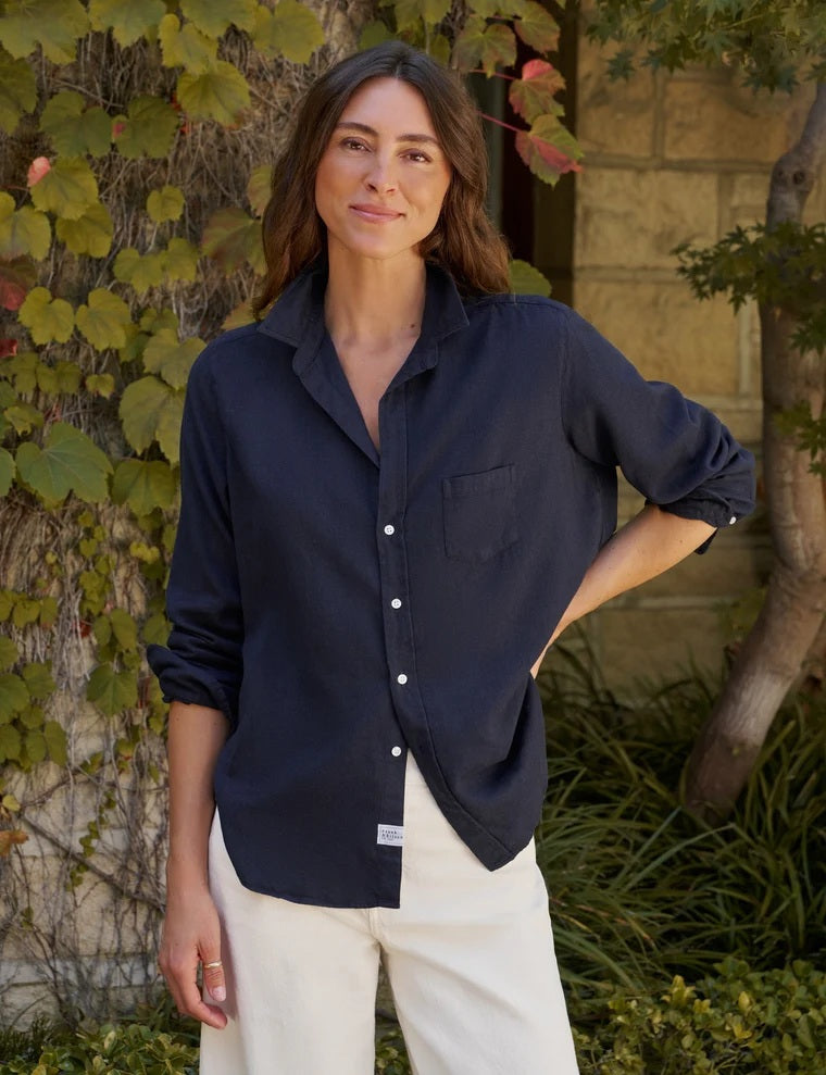 A woman stands barefoot indoors, smiling with her hands in her pockets, dressed in the FRANK & EILEEN EILEEN Relaxed Button Up Shirt in Italian Cashmere Touch Navy and cream wide-leg pants. Sunlight streams through sheer curtains, casting soft shadows across the floor.