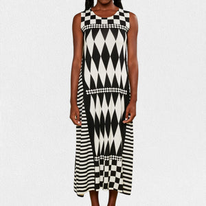 A person is wearing the IN BED WITH YOU - LONG KNIT DRESS featuring a sleeveless, knee-length design and a striking geometric pattern. The dress, from the Italian brand IN BED WITH YOU by CLAUDIOLAI SRL, showcases alternating squares and circles in vibrant prints of red, yellow, purple, and white set against a plain white textured background.