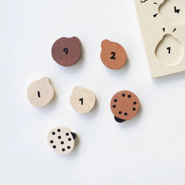 The WEE GALLERY - LADYBUGS COUNT TO 10 TRAY PUZZLE by WEE GALLERY is a wooden puzzle board featuring ladybug-shaped pieces in varying shades from light beige to dark brown. This educational puzzle helps teach counting skills and depicts leaves and twigs alongside the words "Count to Ten" on the left side. Each ladybug is adorned with black dots on their backs to represent number concepts.