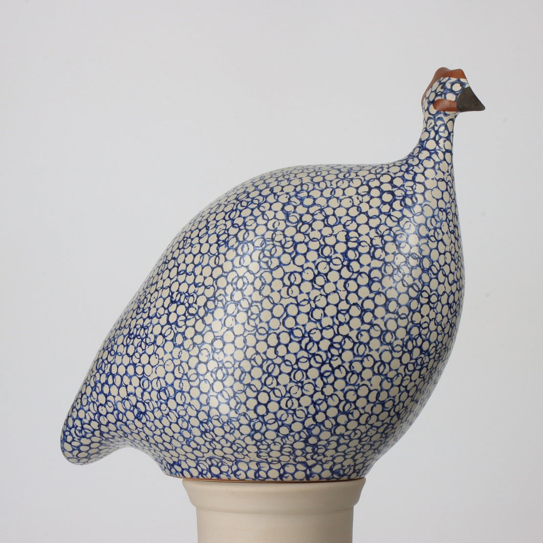 The LES CERAMIQUES DE LUSSAN - LARGE CERAMIC FRENCH GUINEA IN WHITE SPOTTED COBALT MATTE, produced by LES CERAMIQUES DE LUSSAN, is a ceramic sculpture of a guinea fowl. It features an intricate blue and white polka dot pattern on its body and is crafted at our French brood ceramic workshop. The bird, which has a small beak and head with brown accents, is perched on a pedestal against a plain white background.