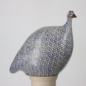The LES CERAMIQUES DE LUSSAN - LARGE CERAMIC FRENCH GUINEA IN WHITE SPOTTED COBALT MATTE, produced by LES CERAMIQUES DE LUSSAN, is a ceramic sculpture of a guinea fowl. It features an intricate blue and white polka dot pattern on its body and is crafted at our French brood ceramic workshop. The bird, which has a small beak and head with brown accents, is perched on a pedestal against a plain white background.