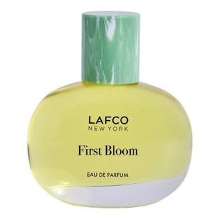 A box of LAFCO - FINE FRAGRANCE, FIRST BLOOM 50ML stands among white flowers and green vines against a pale beige background.