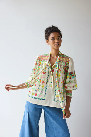 A woman with curly hair looks over her shoulder, wearing the HOPE FOR FLOWERS Smocked Tie Neck Blouse in Custard Floral—a sustainable, cream-colored top with vibrant florals from HOPE FOR FLOWERS, set against a plain, softly lit background.