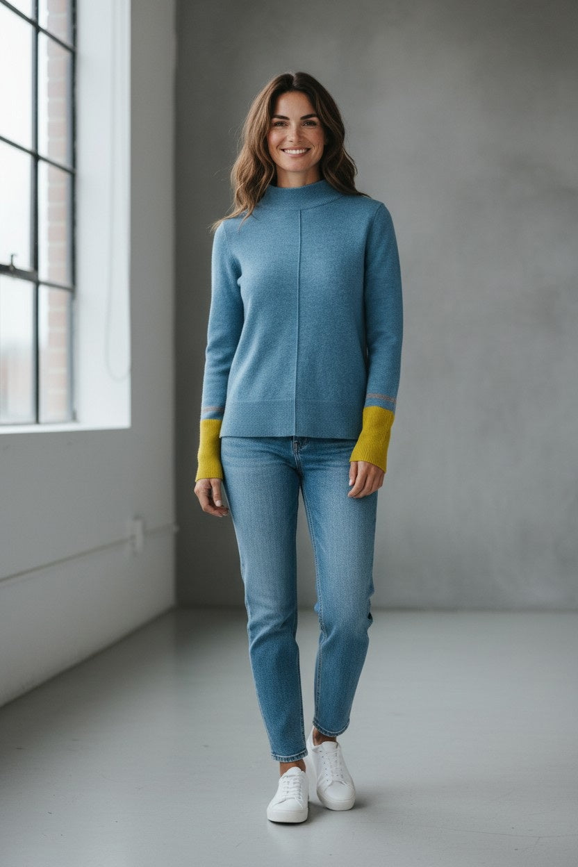A woman stands indoors by a window, smiling with long brown hair, against a plain, neutral-colored wall. She is wearing the KINROSS Colorplay Cashmere Crew Neck Sweater in Loch Multi—a blue cashmere pullover from KINROSS featuring a classic crew neckline and colorblock yellow ribbed cuffs—paired with blue jeans and white sneakers.