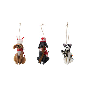 Three HANDMADE FELT DOG ORNAMENTS by CREATIVE COOP sit on a white surface: one wears a red-and-white hat and scarf, another has a black scarf with lights, and the third sports a red plaid hat and scarf. All have string loops for hanging.