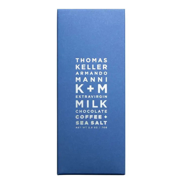 A rectangular blue chocolate bar wrapper with white text that reads: &quot;Thomas Keller Armando Manni K+M Extravirgin Milk Chocolate Coffee + Sea Salt. Net Wt 2.5 oz / 70g&quot;.
