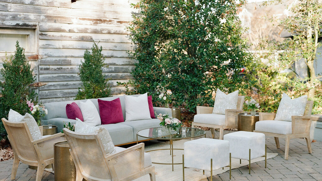 A cozy outdoor seating area features a light gray sofa with pink and white pillows, surrounded by wooden chairs, white ottomans, gold side tables, and a glass coffee table, set against greenery and a rustic wooden wall.