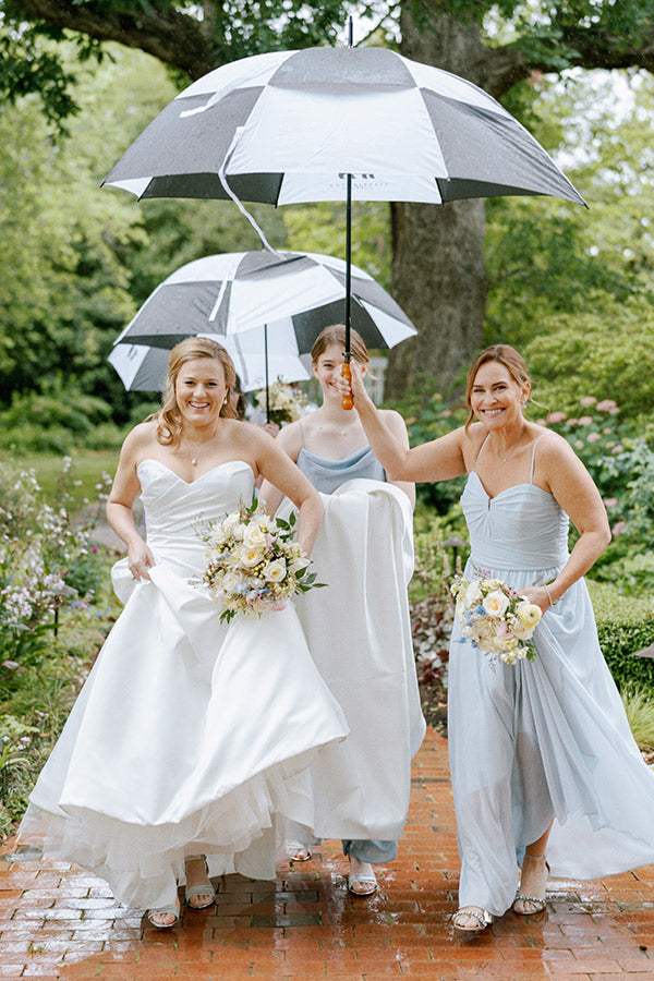 Three women in pastel dresses, holding bouquets and large black-and-white umbrellas, walk on a wet brick path in a lush garden, smiling and appearing joyful despite the rain.