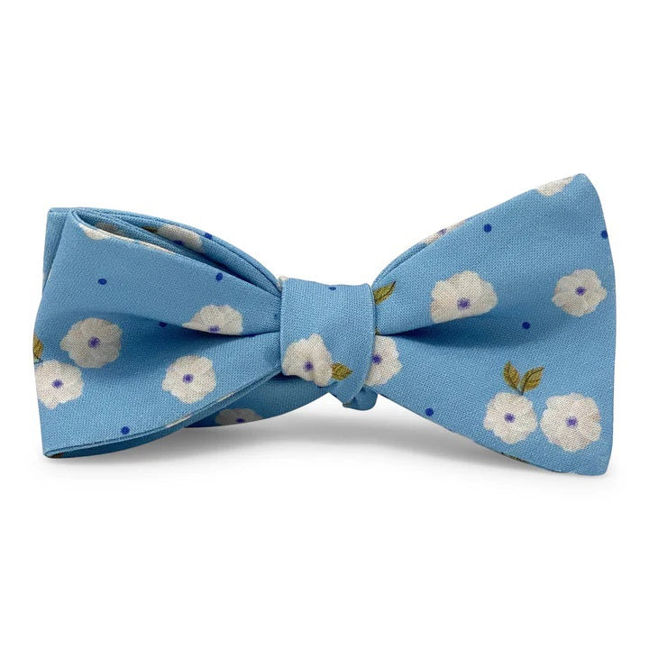 The COLLARED GREENS - COROLLA CAROLINA COTTON BOW TIE IN BLUE by BIRD DOG BAY features a light blue background with small white flowers, green leaves, and scattered blue dots in a handmade design.