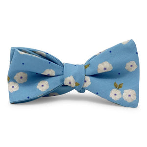 The COLLARED GREENS - COROLLA CAROLINA COTTON BOW TIE IN BLUE by BIRD DOG BAY features a light blue background with small white flowers, green leaves, and scattered blue dots in a handmade design.