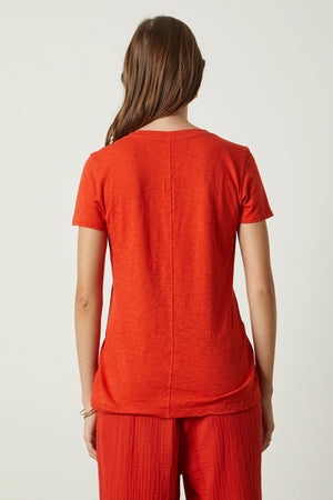 A person with shoulder-length brown hair is standing with their back to the camera. They are wearing a VELVET BY GRAHAM & SPENCER's LILITH V NECK SLUB COTTON TEE in blue and light blue jeans. The background is a plain, light-colored wall.