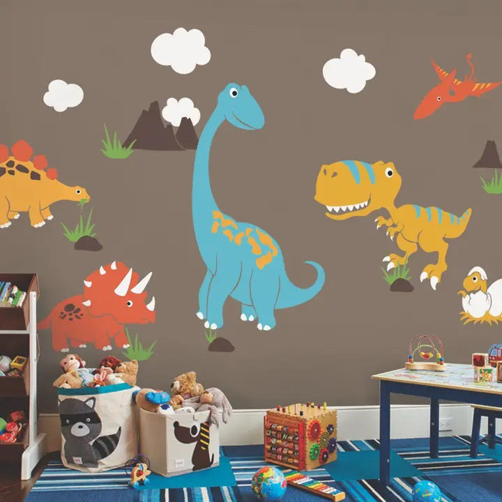 A kids' playroom featuring a brown wall adorned with SIMPLE SHAPES - DINOSAUR REMOVEABLE WALL DECALS by SIMPLE SHAPES. The room, dubbed Dino Paradise, has a blue table, various toys like blocks, a ball, and stuffed animals in a storage bin. Stickers of a blue brachiosaurus, yellow T-Rex, and other cartoon dinosaurs brighten the space.