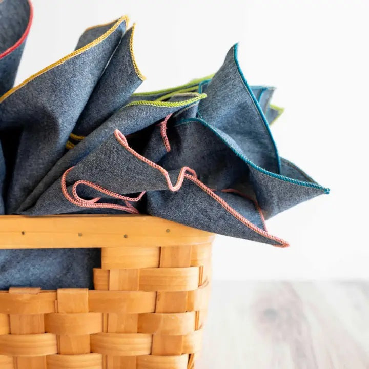 A stack of DOT AND ARMY - CHAMBRAY DINNER NAPKINS rests on a light wooden surface, topped with a sprig of yellow flowers and green leaves.