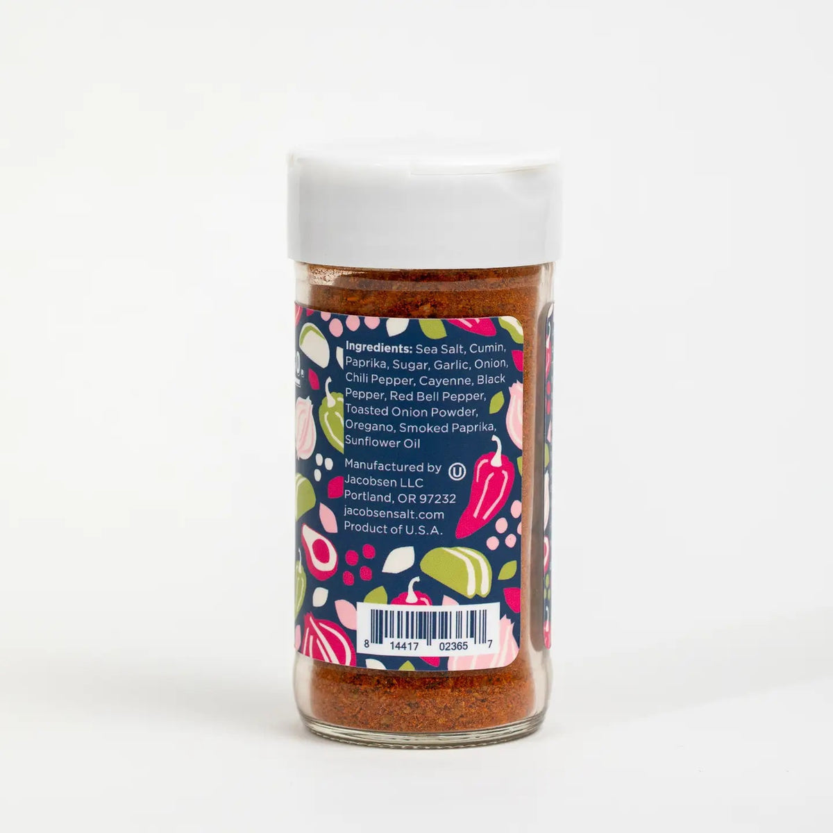 A jar of JACOBSEN SALT - TACO SEASONING from JACOBSEN SALT CO. rests on a kitchen shelf, alongside a white container, wooden utensils, a glass jar with a yellow lid, and a mason jar filled with honey.