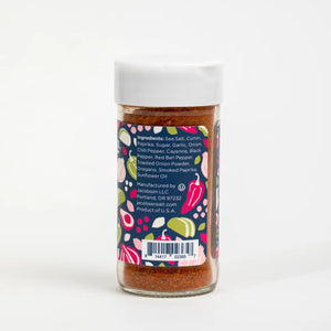 A jar of JACOBSEN SALT - TACO SEASONING from JACOBSEN SALT CO. rests on a kitchen shelf, alongside a white container, wooden utensils, a glass jar with a yellow lid, and a mason jar filled with honey.