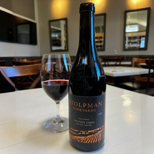 A bottle of STOLPMAN ‘HILLTOPS’ SYRAH 2018 from WINE: WINEBOW, adorned with a black label featuring vineyard graphics and orange text, exemplifies the elegance of California Syrah.