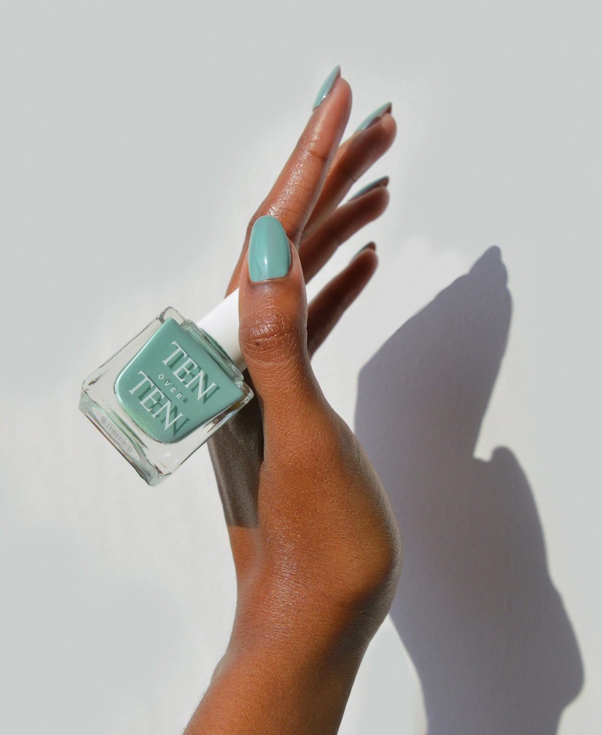 A glass bottle of TENOVERTEN - NAIL POLISH from the brand TEN OVER TEN, featuring a white cap and a dusty pink nail color. The front label displays "TEN OVER TEN" in white uppercase letters. This vegan, eight-free formula ensures both beauty and peace of mind. The background is plain and light-colored.