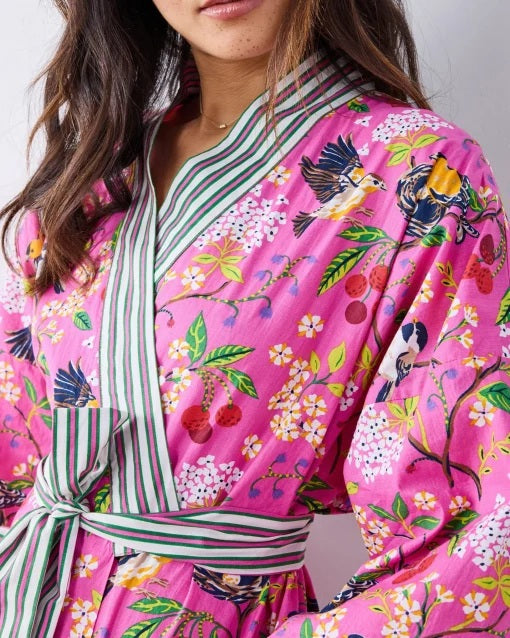 A person wearing a PRINTFRESH - BIRD & BLOSSOM LONG ROBE IN FUSCHIA, a long organic cotton robe with a bird and blossom pattern and green and white striped trim.