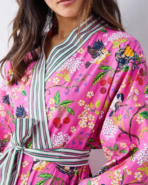 A person wearing a PRINTFRESH - BIRD & BLOSSOM LONG ROBE IN FUSCHIA, a long organic cotton robe with a bird and blossom pattern and green and white striped trim.
