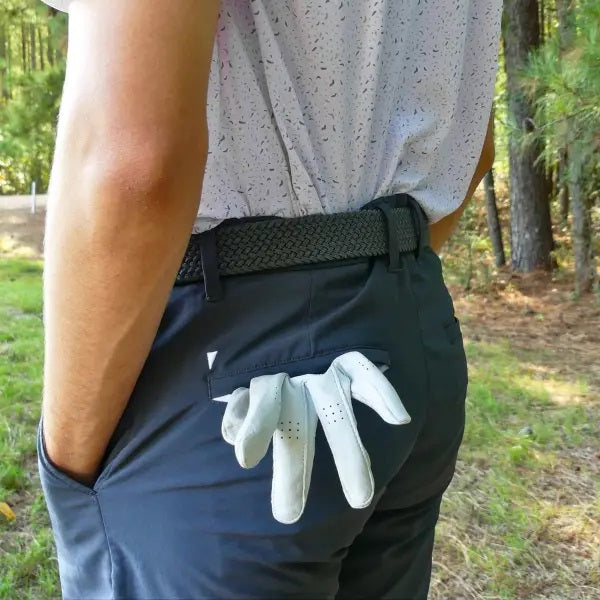A person stands outdoors with hands in pockets, wearing a white short-sleeve button-up shirt, blue shorts, and the ROOSTAS - SCOTTSDALE WOVEN STRETCH BELT by ROOSTAS. Green foliage can be seen in the background.