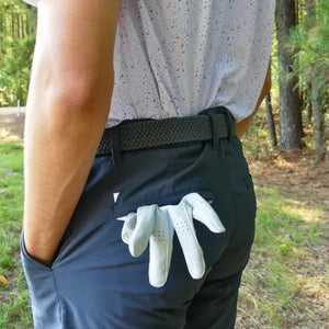A person stands outdoors with hands in pockets, wearing a white short-sleeve button-up shirt, blue shorts, and the ROOSTAS - SCOTTSDALE WOVEN STRETCH BELT by ROOSTAS. Green foliage can be seen in the background.