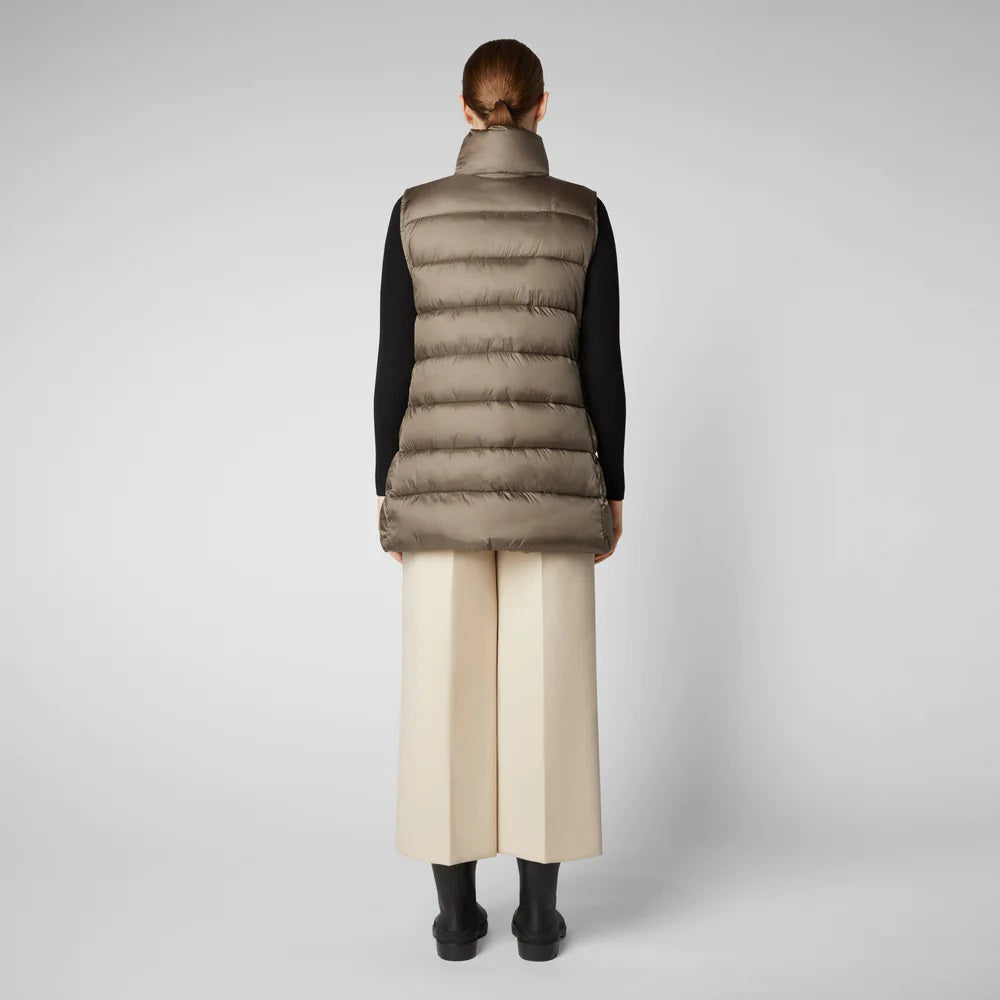 A person wearing a black cap, sunglasses, the SAVE THE DUCK - CORAL PUFFER VEST with animal-free down from SAVE THE DUCK over a black long-sleeve top, beige wide-legged pants, and black ankle boots stands against a plain background. The outfit appears to be styled for colder weather.