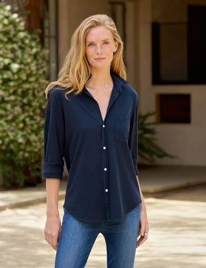 A woman stands in a minimal room, smiling with one hand in her pocket, wearing the FRANK & EILEEN - Waverley Classic Button Up Tee in Vintage Jersey Classic Navy from the California Heritage Collection, paired with blue jeans and brown ankle boots. A large window to the left fills the space with natural light.