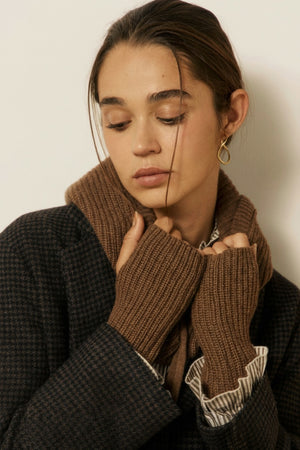 A woman in a dark textured coat and brown knit scarf gazes thoughtfully downward, her hands near her face showing off the elegant HARTFORD - MESSIA Fingerless Mittens in Cognac. A gold teardrop earring completes her ensemble of timeless autumn-winter accessories.
