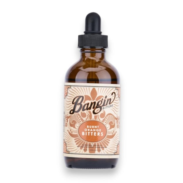 A brown glass dropper bottle labeled “YES COCKTAIL CO - Bangin’ Bitters Burnt Orange” features an ornate, vintage-style design in orange and cream—ideal for adding a zesty twist to craft cocktails from YES COCKTAIL CO.
