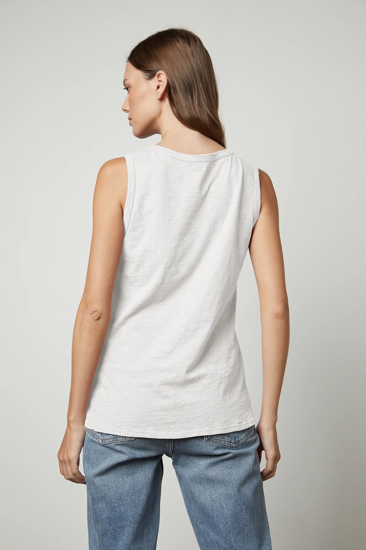 A person with long, brown hair is wearing the VELVET - TAURUS TANK IN COTTON SLUB by VELVET BY GRAHAM &amp; SPENCER and blue jeans while facing the camera. Their right hand is resting on their thigh, and their left hand is visible by their side. The plain, light-colored background complements the tomboy-inspired look.
