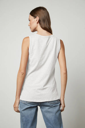 A person with long, brown hair is wearing the VELVET - TAURUS TANK IN COTTON SLUB by VELVET BY GRAHAM & SPENCER and blue jeans while facing the camera. Their right hand is resting on their thigh, and their left hand is visible by their side. The plain, light-colored background complements the tomboy-inspired look.