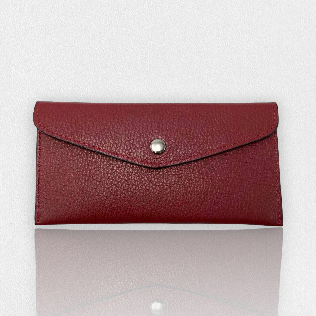 The LISA LIVI LEATHER ENVELOPE WALLET, a stylish money holder, boasts a red textured design with a flap and silver snap button closure set against a plain white background. The bottom edge of the wallet displays a subtle reflection on the surface.