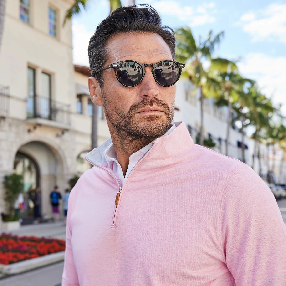A man with short dark hair and a beard wears the COLLARS AND CO PALM PIMA BLEND QUARTER ZIP IN PINK, holding sunglasses in one hand. He stands outdoors by a building with arches, palm trees, and flowerbeds.