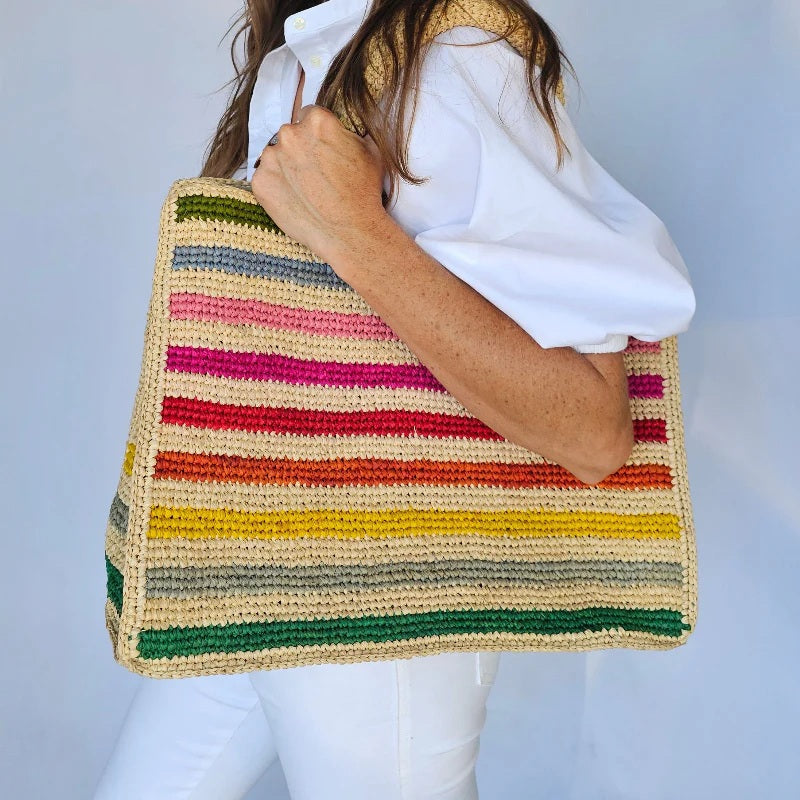 A woman in a white outfit holds the BALDIZ - MAE Raffia Tote in Rainbow, a sustainable handmade bag with colorful stripes by BALDIZ, adding charm to any look against a plain background.