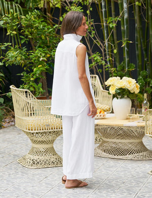 A woman smiles outdoors on a tiled patio, hands in pockets, wearing the FRANK & EILEEN Finley Sleeveless Layering Button Up Shirt in Washed Linen White. Wicker chairs, a flower-topped table, and greenery complete the scene.