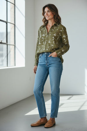 A woman stands in a sunlit room wearing the HARTFORD - CHORUS POPOVER SHIRT IN KHAKI by HARTFORD, paired with blue jeans and brown loafers, smiling with her hands in her pockets.