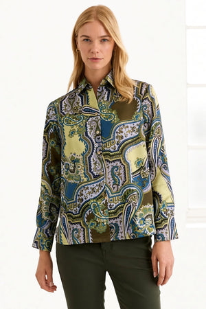 A woman with blonde hair wears the HINSON WU Margot Long Sleeve Paisley Shirt, a colorful button-up featuring green, blue, and yellow paisley patterns and crafted from luxurious Italian polyester for an elegant drape and silky feel. She pairs it with dark green pants and stands against a neutral background.